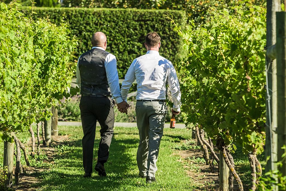 Groom and groomsman walk hand in hand in the vines at Cossars Wineshed Wedding Venue captured by Christchurch wedding photographer Oz