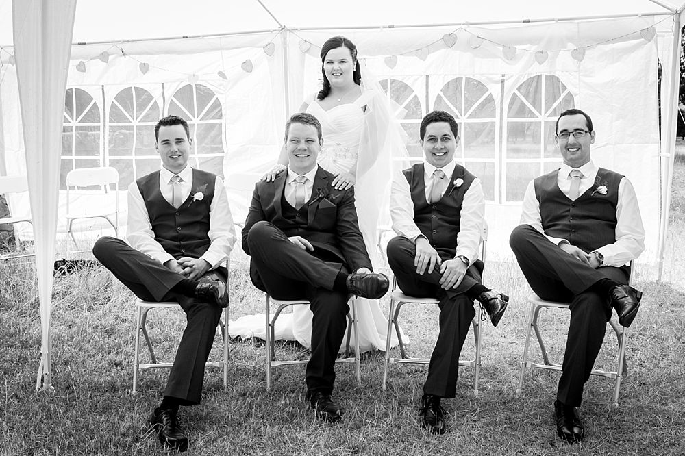 Groom and groomsmen pose for a photo while siting on chairs with bride standing behind the groom